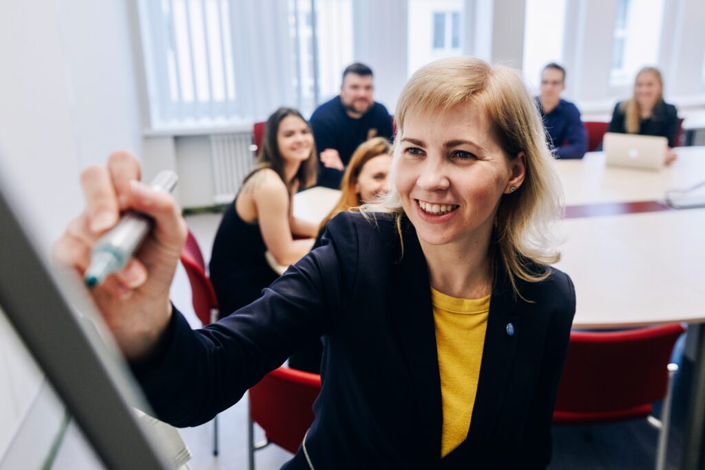 Fotol on inimesed seminarilaua ümber, esiplaanil naine, kes kirjutab midagi tahvlile.